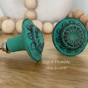 May include: Two teal blue cabinet knobs with a decorative, raised, black metal design. The knobs are made of metal and have a distressed finish. Hip n Humble Hardware.