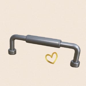 May include: A silver-coloured, bent metal handle with a cylindrical centre section and rounded ends. The handle has a right-angle bend on each end. A gold heart graphic is in the lower right corner.