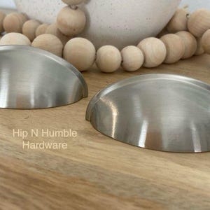 May include: Two silver cabinet knobs with a rounded, slightly domed shape. The knobs are on a light brown wooden surface. Hip N Humble Hardware.