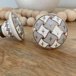 May include: Two ceramic cabinet knobs with a white background and a geometric pattern in shades of grey and gold. The knobs are round and have a slightly raised design.