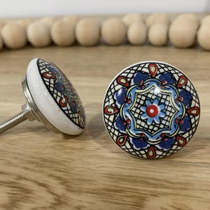 May include: Two white ceramic cabinet knobs with a blue, red and yellow mandala design. The knobs have a silver screw.