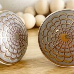 May include: Two white ceramic cabinet knobs with a gold and silver geometric design. The knobs are circular and have a raised design.
