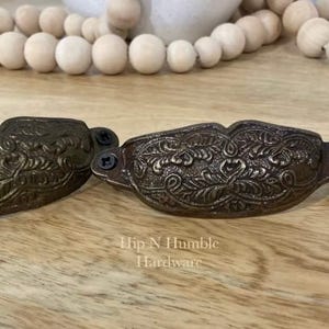 May include: Two antique bronze cabinet pulls with intricate floral and leaf patterns. Each pull is secured with two black screws. The text "Hip N Humble Hardware" is visible on the wooden surface.