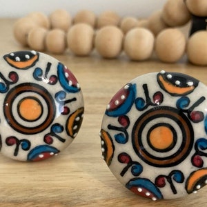 May include: Two white ceramic knobs with a colourful geometric pattern. The pattern features blue, red, orange, brown, and black accents. The knobs have a circular shape and a raised design.