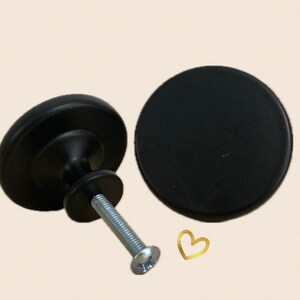 May include: Two black cabinet knobs, one round and one with a raised centre. The round knob is 3.8 cm in diameter. The other knob has a 3.8 cm diameter base and a 2.5 cm diameter centre. Both knobs have a silver screw.