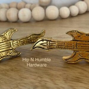 May include: Two gold-coloured guitar-shaped drawer pulls. The pulls have a detailed design and are labelled "Hip N Humble Hardware".