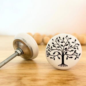 May include: Two white ceramic cabinet knobs with a black tree design. One knob is angled, showing the metal hardware and screw. The other knob is upright, displaying the tree illustration. The knobs are on a wooden surface.