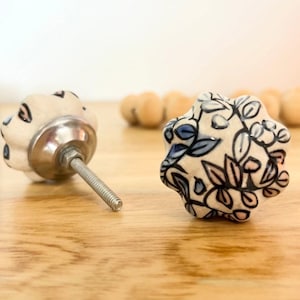 May include: Two ceramic cabinet knobs with floral designs. One is off-white with black and blue accents, the other off-white with black and blue floral patterns. Both have silver metal hardware. They are set against a light wood surface.