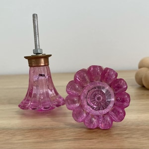 May include: Two pink glass cabinet knobs. One knob is a bell shape with a metal screw. The other knob is a flower shape with multiple petals.