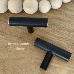 May include: Two black T-shaped cabinet pulls with gold accents. The pulls are on a wooden surface. Hip N Humble Hardware is written in white text on the surface.