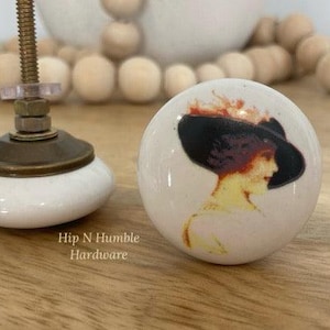 May include: White ceramic knob featuring a vintage illustration of a woman in a black hat. The knob has a brass base and a screw for attaching it to a drawer or cabinet.