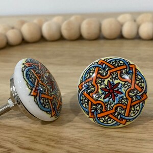 May include: Two ceramic cabinet knobs with a colourful, intricate design. The knobs are white with a yellow, orange, blue, and red pattern. The knobs have a silver metal screw.