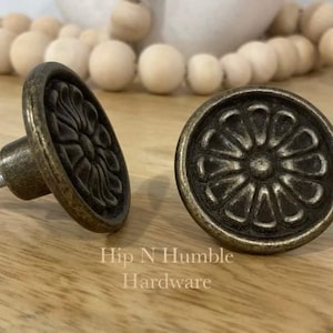 May include: Two antique brass cabinet knobs with a floral design. The knobs are round and have a raised, detailed flower pattern.