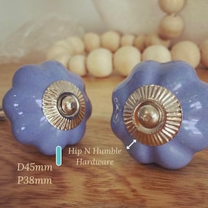 May include: Two lavender ceramic cabinet knobs with a gold metal accent. The knobs are 4.5 cm in diameter and have a 3.8 cm post projection. Hip N Humble Hardware.