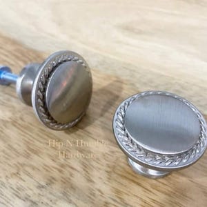 May include: Two silver cabinet knobs with a rope design around the edge. The knobs are round and have a smooth, brushed finish.