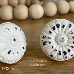May include: Two white ceramic cabinet knobs with a black distressed finish. The knobs have a raised, circular design with a smaller circle in the centre. The text "Hip N Humble Hardware" and "D33mm" are visible below the knobs.