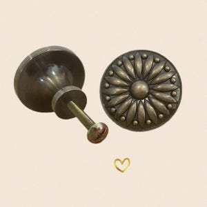May include: Two antique brass cabinet knobs. One knob is round with a raised floral design. The other knob is round with a smooth surface and a screw.