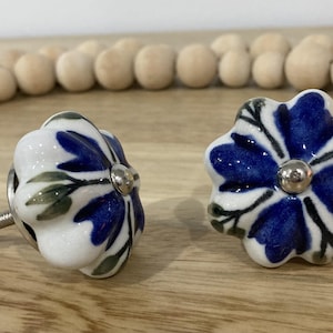 May include: Two ceramic flower-shaped cabinet knobs. The knobs have a white base with blue petals and green leaves. Each knob has a silver metal centre and a screw for installation. Decorative hardware.