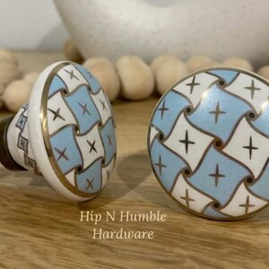 May include: Two ceramic cabinet knobs with a blue and white geometric pattern and gold accents. The knobs are round and have a raised design. The text "Hip N Humble Hardware" is visible below the knobs.