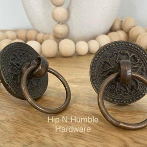 May include: Two antique-style cabinet knobs with ornate, circular, brass-coloured plates and ring pulls. The plates have a detailed floral design.