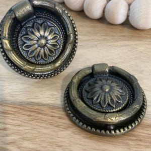 May include: Two antique gold-toned cabinet knobs with ornate floral designs. The knobs have a ring pull and a decorative border.