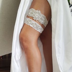 May include: A white bridal garter with a delicate lace design and a large rhinestone embellishment. The garter is worn on the leg of a person wearing a white dress.