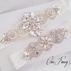 Puede incluir: Dos ligas de encaje blanco con adornos plateados. Una liga presenta un gran dise&ntilde;o floral con pedrer&iacute;a y perlas. La otra liga tiene un dise&ntilde;o floral m&aacute;s peque&ntilde;o con pedrer&iacute;a. El texto "One Fancy Day" es visible en la esquina inferior derecha de la imagen.