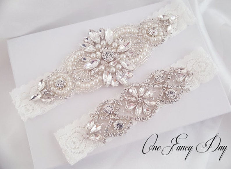 Puede incluir: Dos ligas de encaje blanco con adornos plateados. Una liga presenta un gran dise&ntilde;o floral con pedrer&iacute;a y perlas. La otra liga tiene un dise&ntilde;o floral m&aacute;s peque&ntilde;o con pedrer&iacute;a. El texto "One Fancy Day" es visible en la esquina inferior derecha de la imagen.