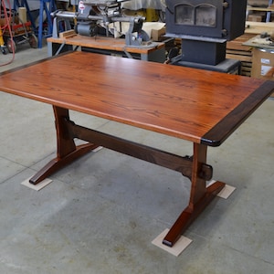 May include: A solid wood table with a dark brown border around the edge of the top. The table has a trestle base with a single stretcher.