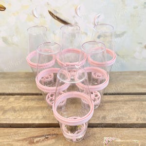 May include: A set of six clear plastic champagne flutes arranged in a triangular formation. Each flute features a light pink circular base with a decorative cutout pattern and a matching light pink rim around the middle of the glass body.