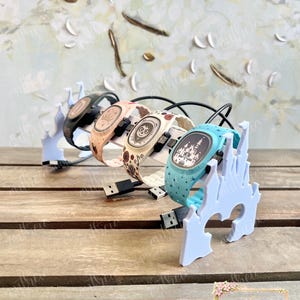 May include: Four digital watches with themed designs and matching light blue charging stands. The watches have black, pink, white, and blue bands. Each watch is connected to a USB charging cable. The background is a light floral design.