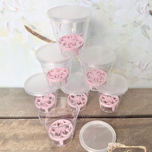May include: A stack of seven clear plastic cups with lids, each containing a light pink, circular, floral-patterned insert at the bottom. One additional lid is placed separately in the foreground. The cups are arranged in a pyramid formation.