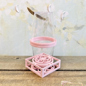 May include: A clear plastic tumbler with a light pink rim and a light pink square base featuring a cutout floral design. The tumbler is inverted and rests on the base, which displays a central circular pattern with radiating petals.