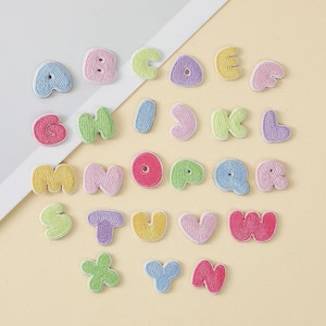 Könnte beinhalten: Eine Sammlung von sechsundzwanzig einzelnen gestickten Alphabet-Buchstaben-Aufnähern in verschiedenen Pastellfarben wie Rosa, Blau, Grün, Gelb und Lila. Jeder Buchstabe hat eine abgerundete Schriftart im Blasenstil mit einer gestickten Randtextur.