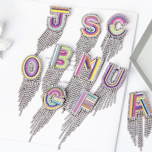 May include: A set of colourful letter earrings with rainbow stripes and crystal fringe. The letters are J, S, C, O, B, M, U, G, F, and A.