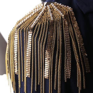 Könnte beinhalten: Ein Schulterstück aus goldener Kette mit einem großen Strasssteinbesatz an der Oberseite. Die Kette besteht aus kleinen, miteinander verbundenen Kreisen.