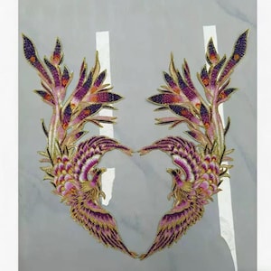 May include: Two embroidered phoenix patches. The patches feature a detailed design with purple, pink, and gold thread. The wings are spread, and the overall shape is symmetrical. The background is a light gray color.