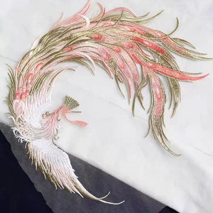 Puede incluir: Una aplicación bordada de fénix rosa y dorada con detalles intrincados. El fénix se representa con las alas extendidas, creando un diseño dramático y elegante.