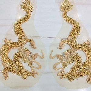 Puede incluir: Dos aplicaciones de dragón bordadas en oro sobre un fondo de tela transparente. Los dragones están detallados con lentejuelas y cuentas.