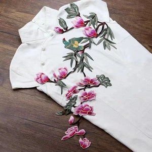 Puede incluir: Una tela blanca con un diseño floral bordado en rosa y verde. El diseño presenta un pájaro posado en una rama con flores rosas y hojas verdes.