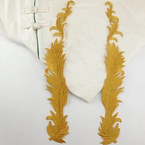 Pode incluir: Dois apliques de tecido de cor dourada em forma de penas estilizadas. Os apliques estão sobre um fundo branco.