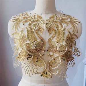 May include: Gold sequined applique featuring two swans with pearl accents. The applique is on a sheer mesh fabric and is perfect for adding a touch of elegance to any garment.