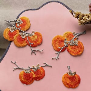 May include: Embroidered orange persimmon fruit patches on gray branches. The patches are in various sizes and arrangements, with detailed stitching and a gradient of orange and red hues. The background is a light pink surface.