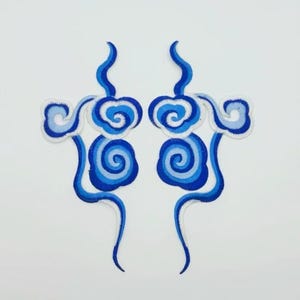 May include: Two blue and white embroidered patches in the shape of stylised clouds. The clouds are layered with different shades of blue and white.