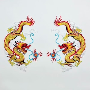 May include: Two embroidered dragon patches. Each dragon is yellow with red and orange accents, and blue and white details. The dragons face each other, with their bodies curved and their heads raised. The patches are on a white background.