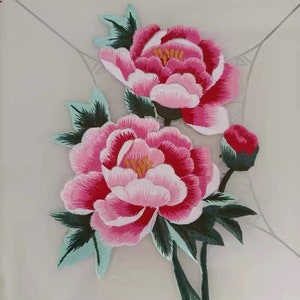 May include: Embroidered floral applique featuring two large pink peonies with white and red accents, green leaves, and a bud. The flowers have yellow centers and are set against a light background. This decorative patch is ideal for fabric crafts.