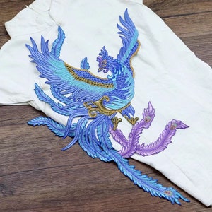 May include: A blue and gold embroidered phoenix applique with purple feathers. The phoenix is a mythical bird that symbolizes rebirth and immortality.