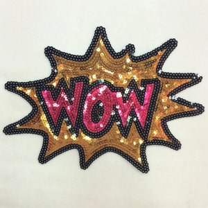 Peut inclure: Un patch thermocollant en sequins dorés et roses en forme de bulle de dialogue avec le mot "WOW" à l'intérieur.