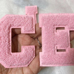 Può includere: Due lettere in feltro rosa, C e H, con un contorno bianco. Le lettere sono cucite insieme e hanno una texture pelosa.