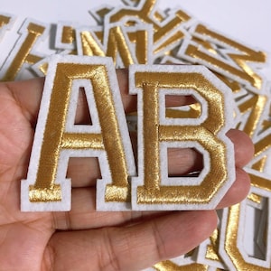 Op de afbeelding: Goud geborduurde letters A en B met een witte rand. De letters zijn op een witte vilt ondergrond.